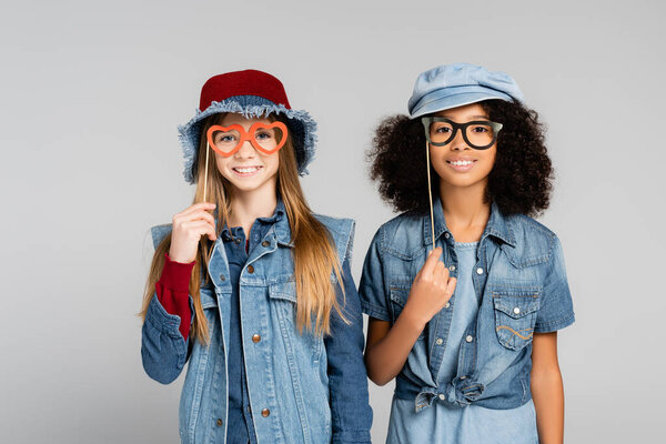 smiling multicultural girls in denim clothes holding party spectacles isolated on grey