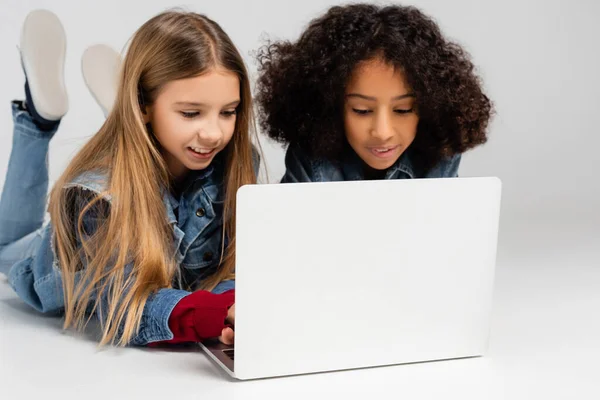 Smiling Trendy Interracial Girls Lying Grey Using Laptop Together — Stock Photo, Image