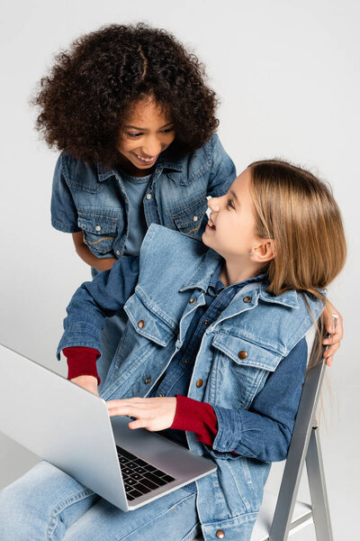 cheerful african american girl in denim clothes near happy friend sitting on chair with laptop isolated on grey