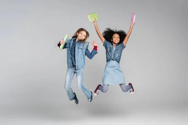 Aufgeregte Multikulturelle Schulmädchen Jeanskleidung Schweben Mit Schulbüchern Auf Grau — Stockfoto