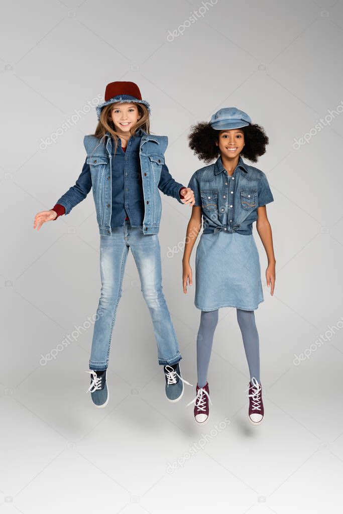 full length view of joyful multicultural girls in trendy denim clothes levitating on grey