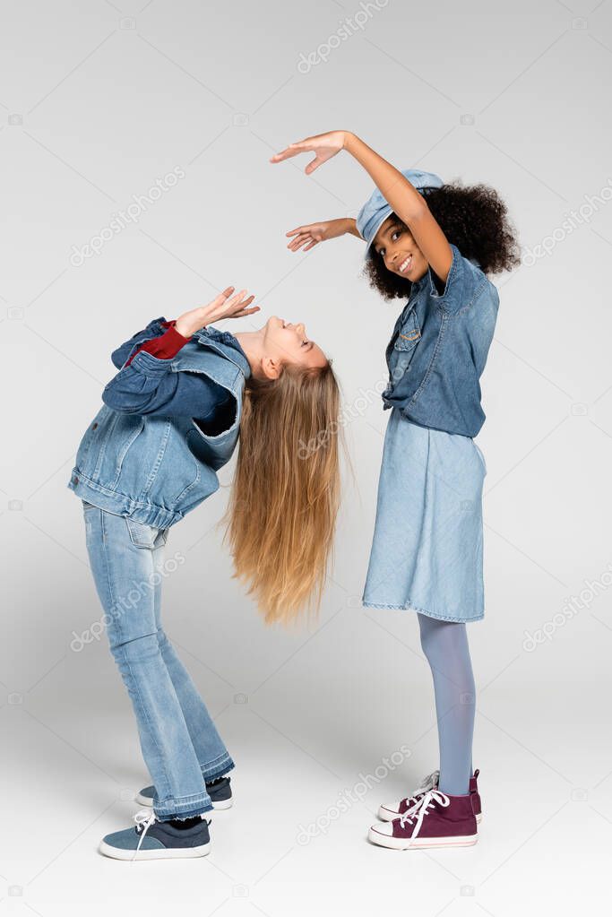 joyful african american girl looking at camera while showing scaring gesture near friend on grey