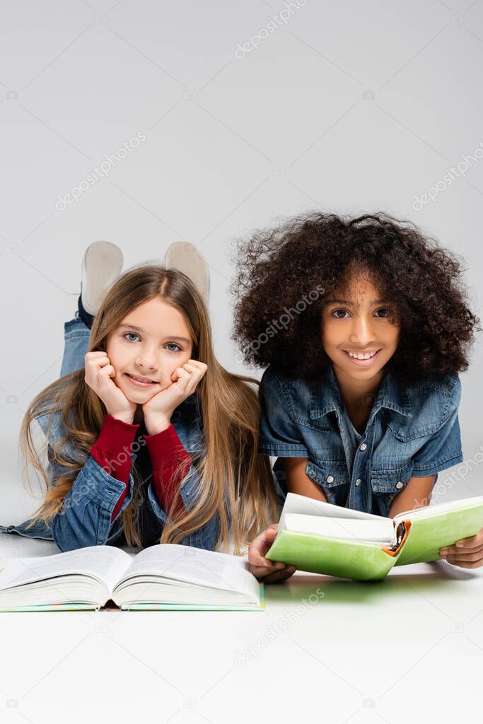 happy multicultural schoolkids smiling at camera while lying with books on grey