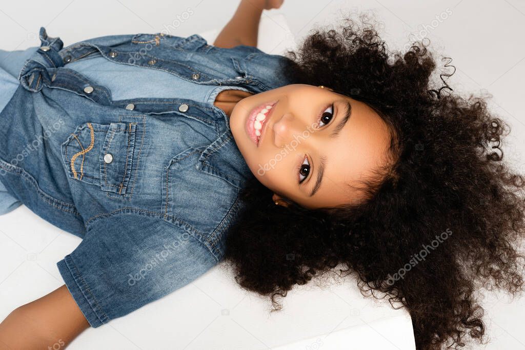 happy african american girl in stylish denim clothes smiling at camera while lying on white surface isolated on grey