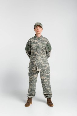 Woman in military uniform and boots looking at camera on grey background  clipart
