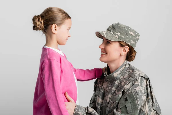 Smiling mother in military clothes hugging daughter isolated on grey