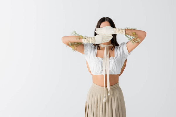 brunette woman covering face with hands in gloves with flowers isolated on white