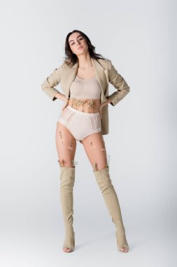 Spor ceketli, çizmeli ve külotlu genç bir kadın. Çiçek açmış, elleri beyaz kalçasında poz veriyor.