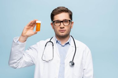 young doctor in glasses and white coat holding bottle with medication isolated on blue clipart