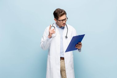 Gözlüklü ve beyaz önlüklü doktor fikir işareti gösteriyor ve mavi panoya bakıyor.