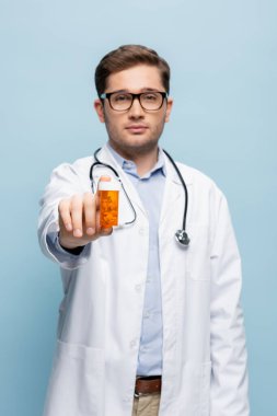 Bulanık gözlüklü genç doktor ve elinde ilaç şişesi olan beyaz önlüklü mavi haplı.