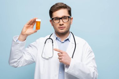 doctor in glasses and white coat pointing with finger at bottle with medication isolated on blue clipart