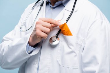 Beyaz önlüklü, steteskoplu doktorun kırpılmış görüntüsü mavi üzerine izole edilmiş ilaç şişesi koyuyor.