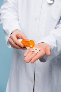 partial view of doctor in white coat holding pills and bottle in hands isolated on blue clipart