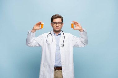 young doctor in glasses and white coat holding bottles with remedy on blue clipart