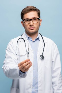 Doktor gözlüklü ve beyaz önlüklü aşı şırıngası taşıyor.