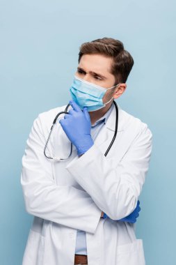 Tıbbi maskeli düşünceli bir doktor ve mavi üzerine steteskop takmış beyaz önlüklü.