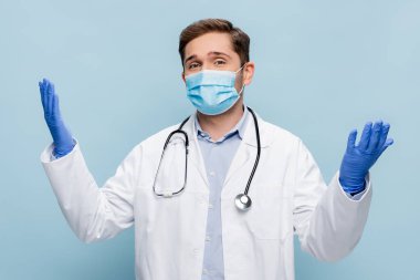 Tıbbi maskeli genç bir doktor ve beyaz önlüklü uzanmış elleri mavi üzerinde duruyor.