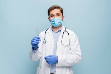 Tıbbi maskeli genç bir doktor ve steteskobu olan beyaz önlüklü mavi şırıngayı tutan.