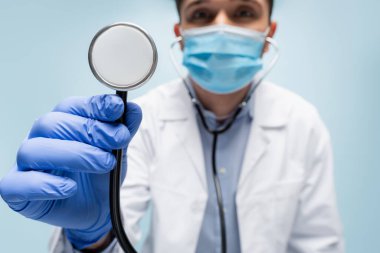 Tıbbi maskeli ve beyaz önlüklü bulanık genç doktor stetoskobu mavi üzerine izole etti.