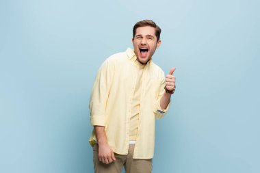 excited young man looking at camera and showing thumb up on blue clipart