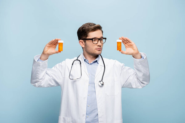 молодой врач в очках и белом халате, держащий бутылки с таблетками на синем