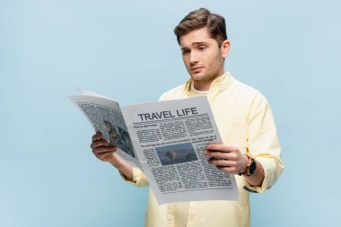 Tişörtlü kuşkucu genç adam mavi üzerine izole edilmiş seyahat gazetesi okuyor.