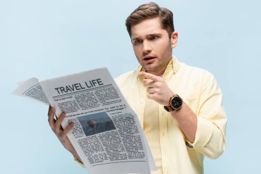 Tişörtlü genç adam mavi üzerine izole edilmiş seyahat gazetesini işaret ediyor.
