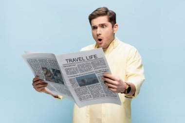 Şok olmuş genç adam mavi üzerine izole edilmiş seyahat gazetesi okuyor.