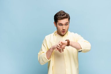 surprised young man in yellow shirt looking at wristwatch on blue clipart
