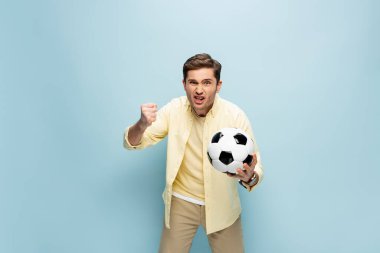 Sarı tişörtlü rahatsız bir adam elinde mavi futbol topu tutuyor.