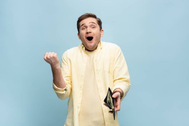 Sarı gömlekli heyecanlı genç adam cüzdanı mavi üzerinde dolarlarla birlikte tutuyor.