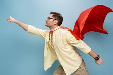 side view of superhero in glasses and red cape standing with outstretched hand on blue clipart