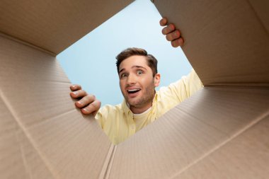 bottom view of curious man looking at camera from carton box isolated on blue clipart