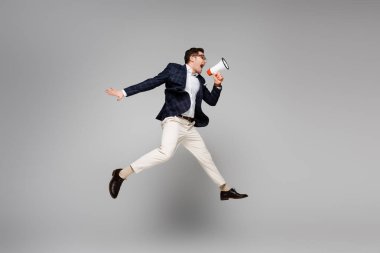 full length of businessman jumping and screaming in megaphone on grey clipart