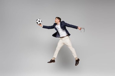 Tam boy gülümseyen iş adamı gözlüğü tutuyor ve gri üzerinde havada süzülürken futbol oynuyor.