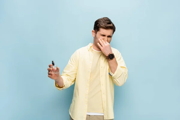 disgusted man in yellow shirt covering nose and holding bottle with perfume on blue