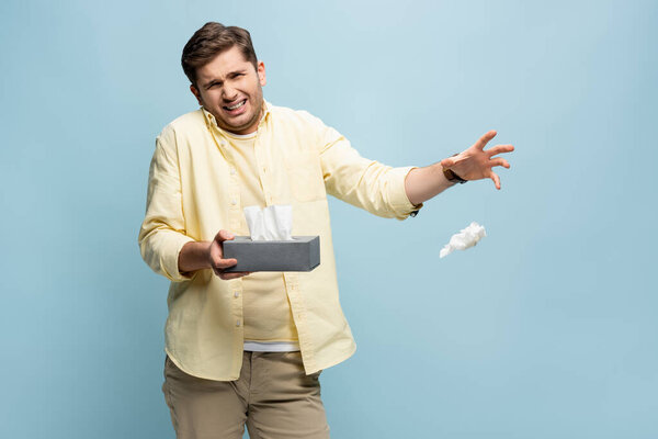 sick man throwing napkin away and holding tissue box isolated on blue