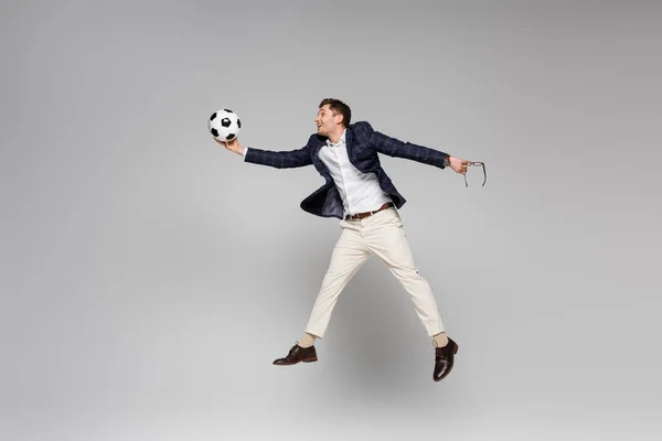full length of smiling businessman holding glasses and playing football while levitating on grey