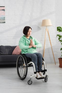 Tekerlekli sandalyedeki kadın dua eden elleriyle evde oturuyor. 