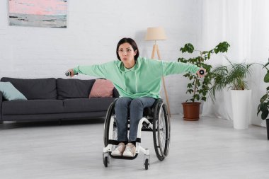 Tekerlekli sandalyedeki kadın oturma odasında dambıllarla çalışıyor. 