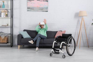 VR kulaklıklı heyecanlı kadın oturma odasında tekerlekli sandalyenin yanında oturuyor. 