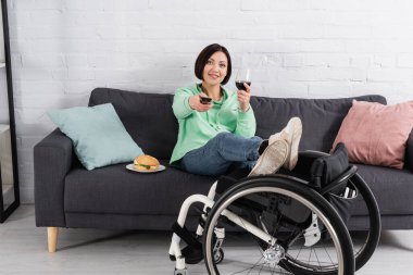 Handicapped woman with remote controller and glass of wine sitting near hamburger and wheelchair at home  clipart
