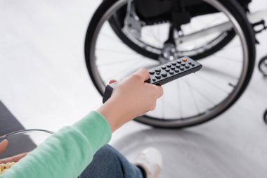 Arka planda tekerlekli sandalyenin yanında uzaktan kumanda ve patlamış mısır tutan bir kadın görüntüsü. 