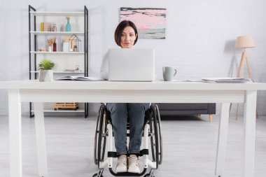Tekerlekli sandalyedeki kadın dizüstü bilgisayarla masa başında çalışıyor. 