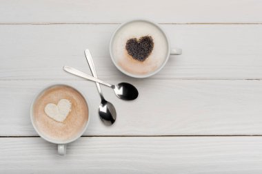 top view of cups with cappuccino and latte near spoons on white wooden surface  clipart