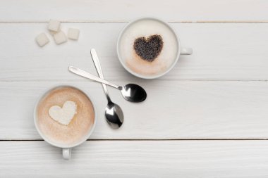 Kapuçinolu fincanların üst görüntüsü ve kaşıkların yanında latte ve beyaz ahşap zemin üzerinde şeker küpleri  