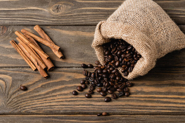 top view of sack bag with roasted coffee beans near cinnamon sticks on wooden surface 