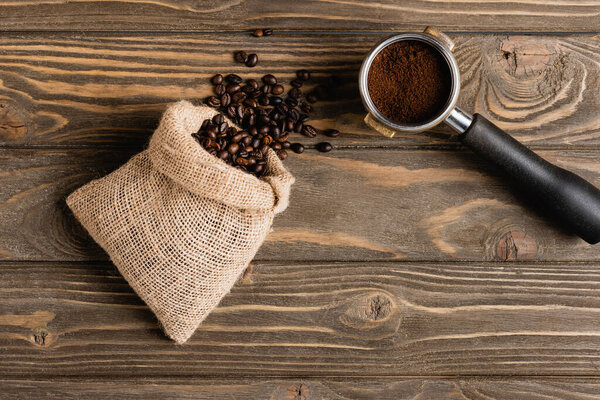 top view of sack bag with beans near metallic portafilter with fresh ground coffee on wooden surface