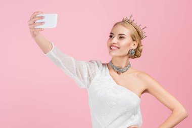 Lüks taclı sarışın kadın pembe renkte izole edilmiş akıllı telefondan selfie çekiyor.